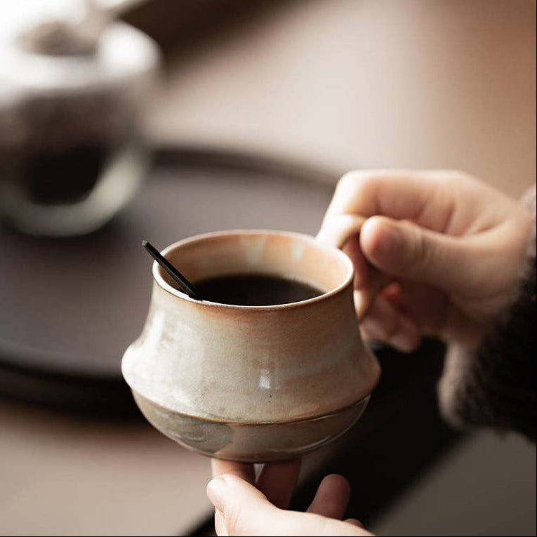 A close-up of a person holding a small handmade ceramic cup filled with black coffee, with a spoon resting inside. The cup has a rustic, earthy beige and brown glaze.